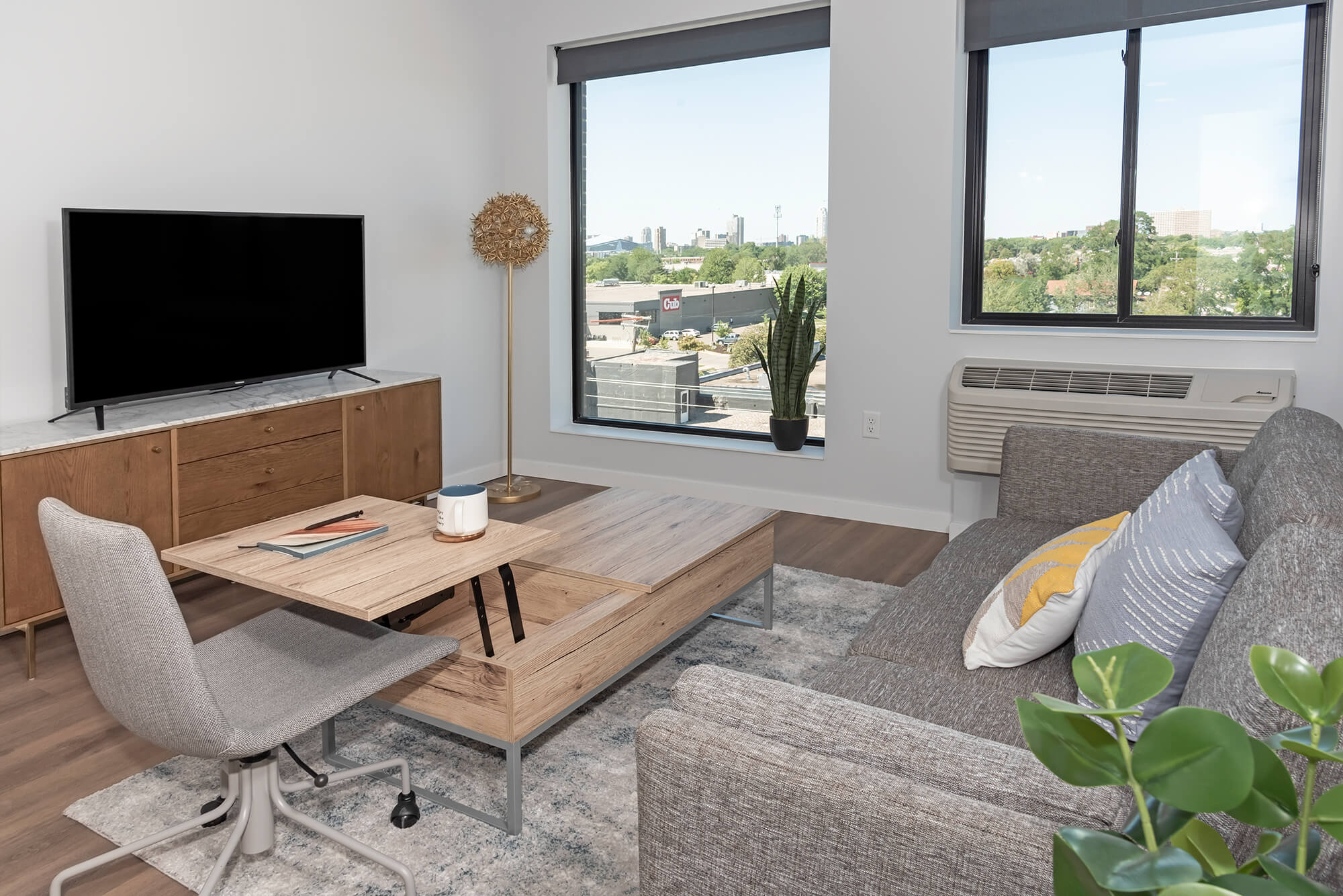 Apartment living room with TV, gray sofa, coffee table, and large windows with city views.