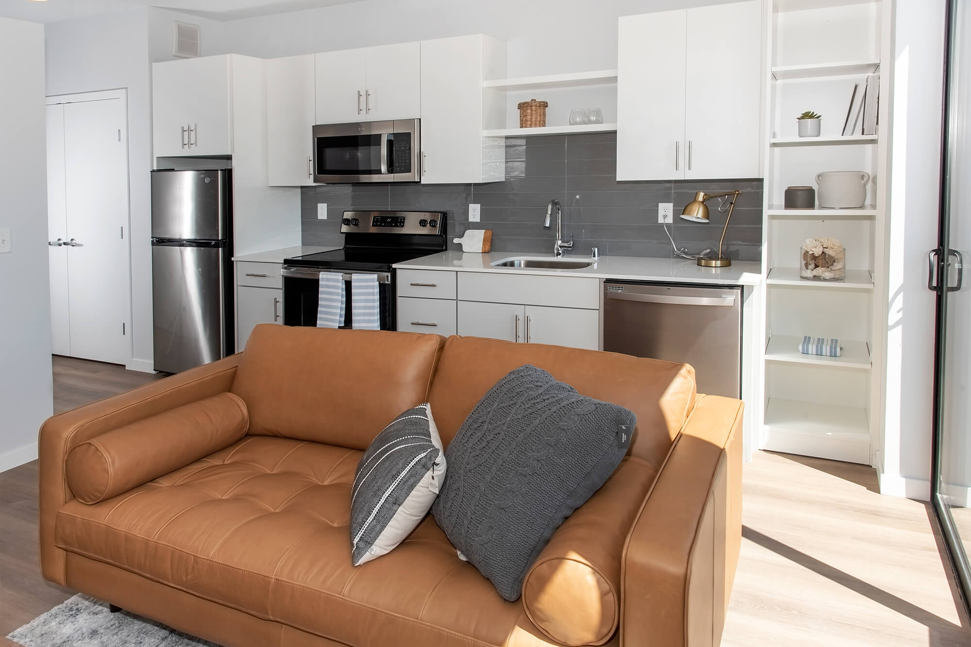 Modern apartment kitchen with white cabinetry, stainless steel appliances, and leather sofa in the foreground.