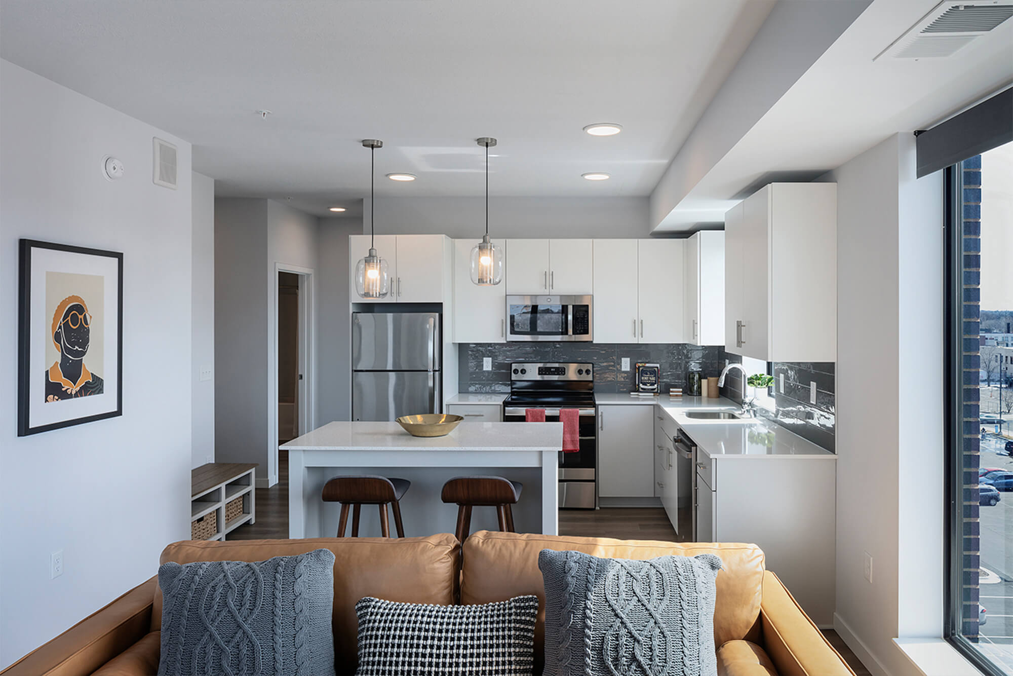 Open-concept apartment living room and kitchen with island seating, stainless steel appliances, and large windows.