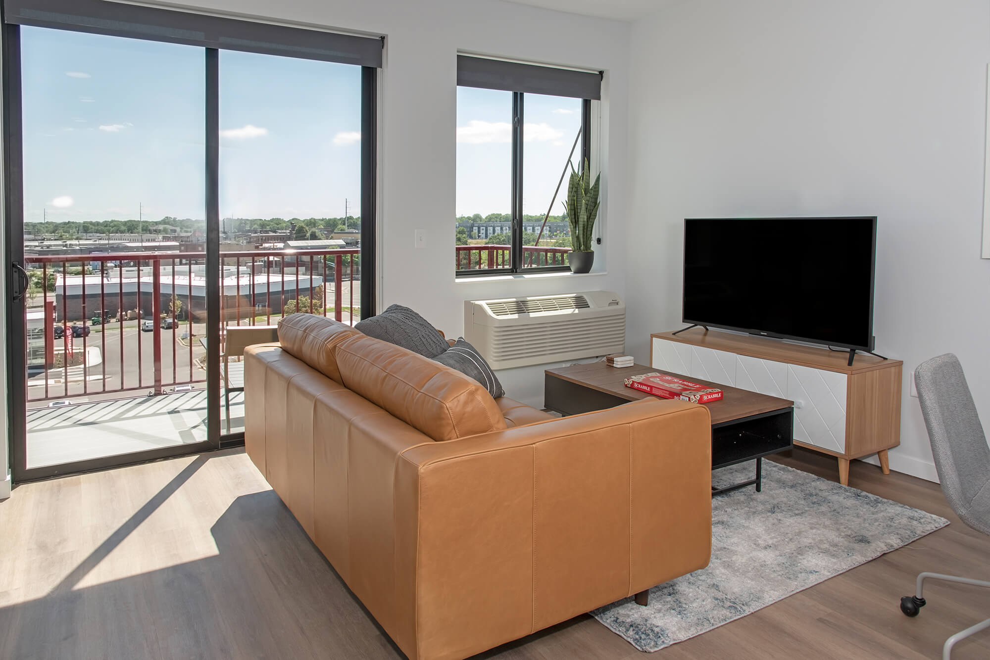 Apartment living room with leather sofa, TV, desk chair, and balcony access with large windows.
